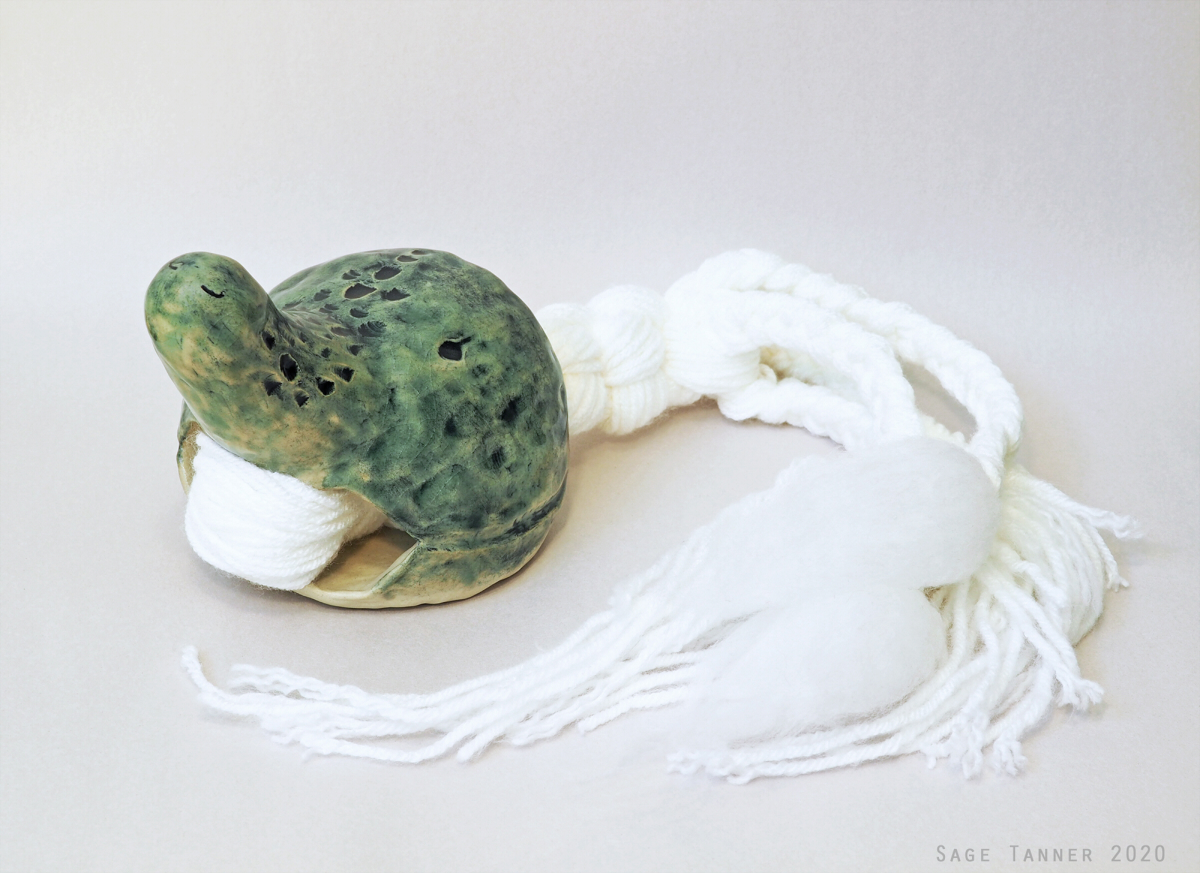 A creature-like doll with a green glazed ceramic body and a white braided yarn tail.